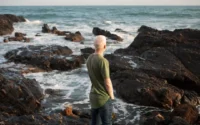 Person standing calmly near a stormy ocean with rays of sunlight breaking through dark