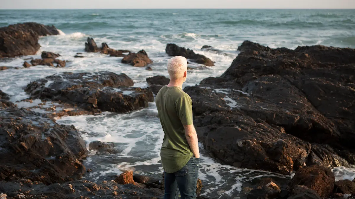 Person standing calmly near a stormy ocean with rays of sunlight breaking through dark