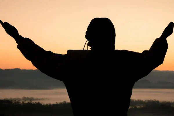 Silhouette of a person praying with hands raised while light shines through darkness, representing spiritual victory in Jesus.