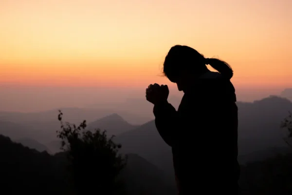 Text: A person praying calmly in a well-lit room while shadows and darkness are blurred and kept at a distance in the background.