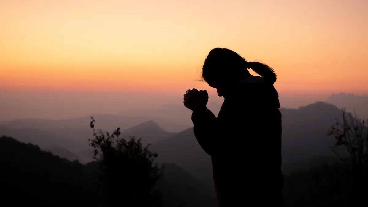 Text: A person praying calmly in a well-lit room while shadows and darkness are blurred and kept at a distance in the background.